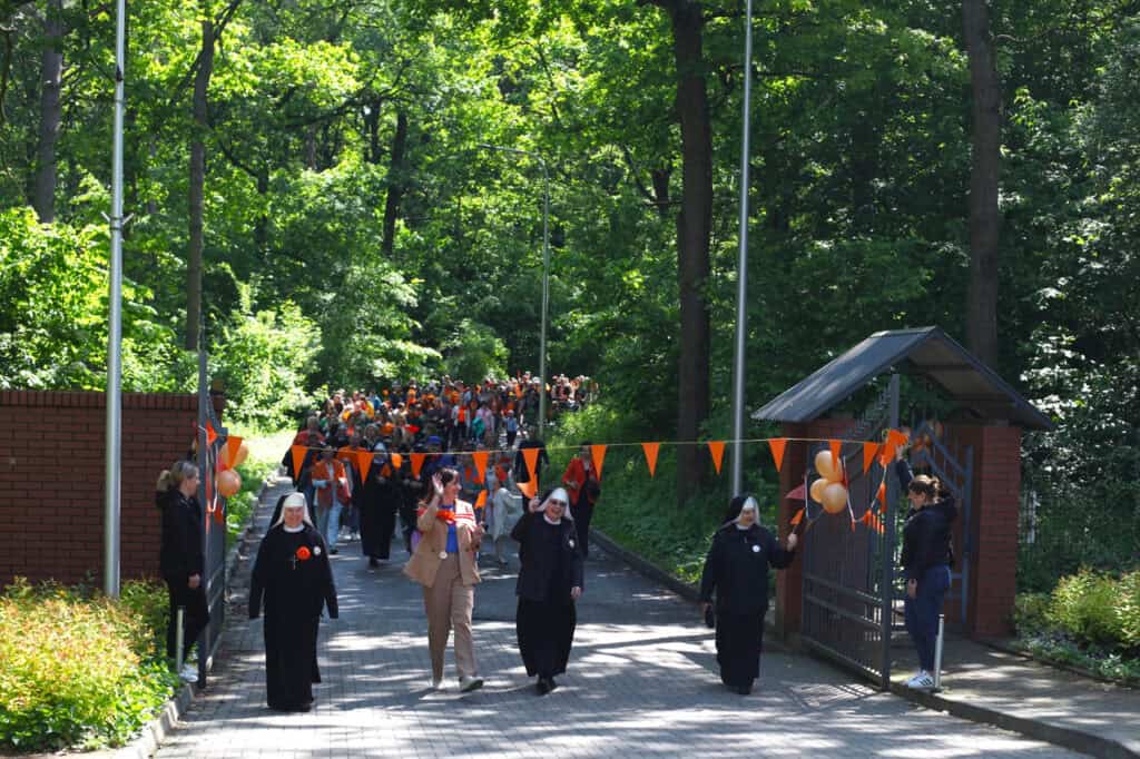 Wejście pierwszych uczestników marszu na teren fundacji. Symboliczne przejście pod pomarańczową wstęgą, na której powiewają pomarańczowe proporce
