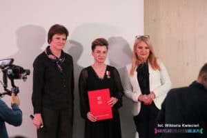 Od lewej stoją: Joanna Olszewska, Karolina Czajka oraz Anna Augustyniak. Kobieta w środku trzyma w rękach czerwoną okładkę z aktem nadania Medalu Prezydenta Miasta Lublin "Zasłużony dla Miasta Lublin"