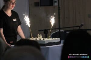 Kelnerka pcha wózek serwisowy, na którym stoi okazały tort z okazji 35-lecia. Ozdobiony jest dwiema świeczkami w kształcie cyfr „3” i „5” oraz płonącymi racami, z których tryskają jasne iskry