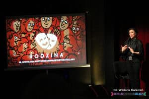 Obok ekranu multimedialnego z planszą zapowiadającą 35. Spotkania Artystów Nieprzetartego Szlaku "Rodzina" stoi Pani Magdalena Gach - tłumacz PJM