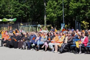 Zgromadzona publiczność siedząca na krzesłach. W pierwszym rzędzie przedstawiciele władz, instytucji, zaproszeni goście. W tle zieleń drzew