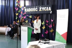 Na tle granatowej kurtyny z kolorowymi motylami i napisem „Radość życia” od lewej: tłumacz języka migowego. Następnie prelegentki: Barbara Sadowska-Burlita (ekspert ds. CIDON) oraz Iwona Podkowa-Nowak (specjalista ds. CIDON) podczas wystąpienia. Po prawej stronie znajduje się baner informacyjny PFRON