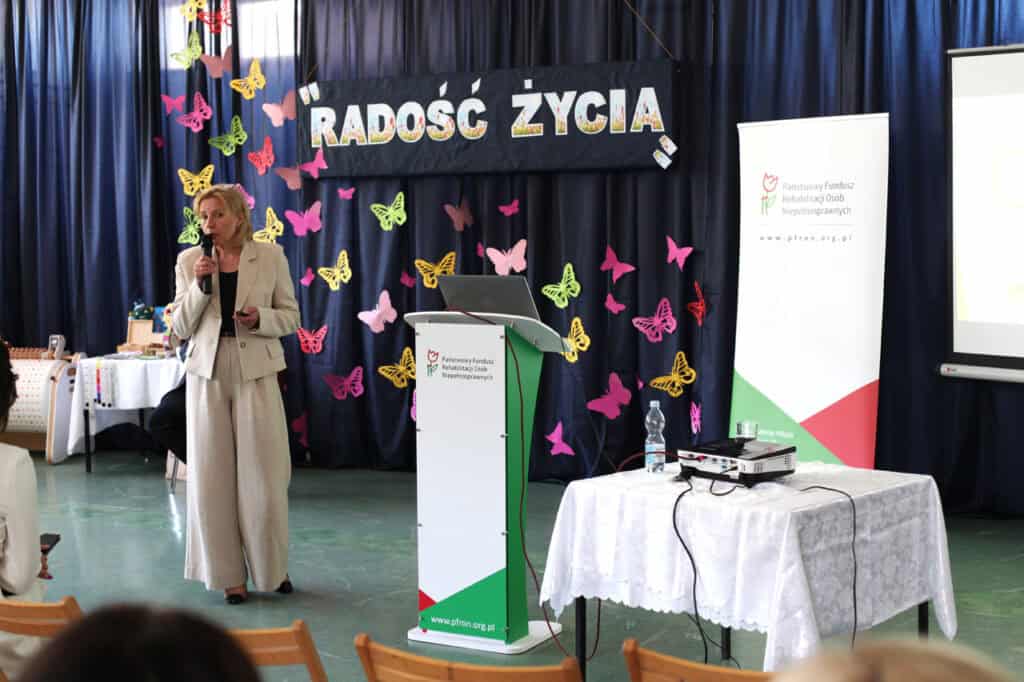 Małgorzata Paprota (kierownik OWiT Świdnik) podczas wystąpienia. Tło stanowi granatowa kurtyna z motylami w różnych kolorach (różowe, zielone, żółte) i napis „Radość życia”