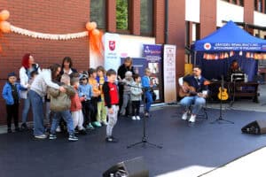 Na scenie dzieci prezentujące utwór muzyczny. Na pierwszym planie solista stojący przed mikrofonem. W prawej części kadru gitarzysta akompaniujący