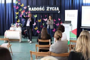 W centralnej części kadru: Jakub Kozik i Paulina Kowalik podczas wykładu przed zebranymi słuchaczami. Po lewej stronie prezentowane urządzenie Musicon. Tuż za nim stolik z modułami do urządzenia