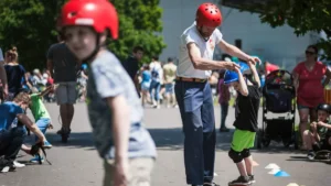 Mężczyzna w czerwonym kasku i białej koszuli pomaga trzymając za ręce młodego chłopca, który również ma na sobie niebieski kask i jeździ na deskorolce. Chłopiec jest skupiony. W tle widoczny jest tłum ludzi, wśród nich dzieci i dorośli, którzy cieszą się rekreacyjnymi aktywnościami.