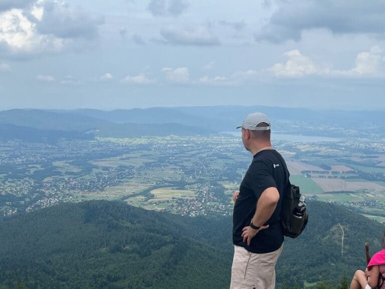 Mężczyzna stoi tyłem do aparatu na punkcie widokowym. Przed nim rozciąga się panorama – poniżej widać gęsty las, niewielkie zabudowania i pola uprawne, a w oddali pasmo górskie