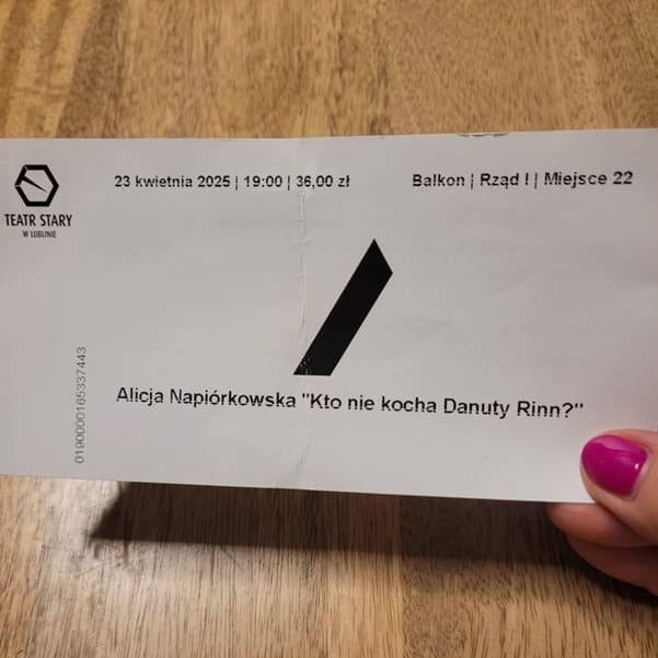 Na tle blatu w kolorze drewna kobieca dłoń z różowym paznokciem trzyma biały bilet wstępu z czarnym nadrukiem