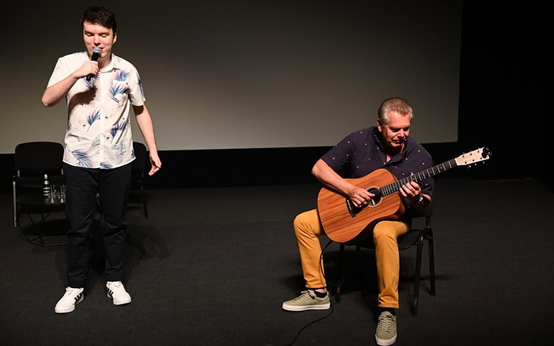 Mężczyzna stoi i śpiewa do mikrofonu, podczas gdy inny siedząc na krześle gra na gitarze