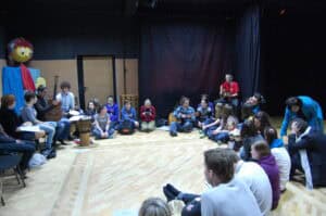 Siedziba Centrum Nieprzetartego Szlaku w Lublinie przy ul. Nowy Świat. W sali teatralnej podczas spotkania po latach, grupa wolontariuszy siedzi w kręgu; niektórzy trzymają instrumenty muzyczne