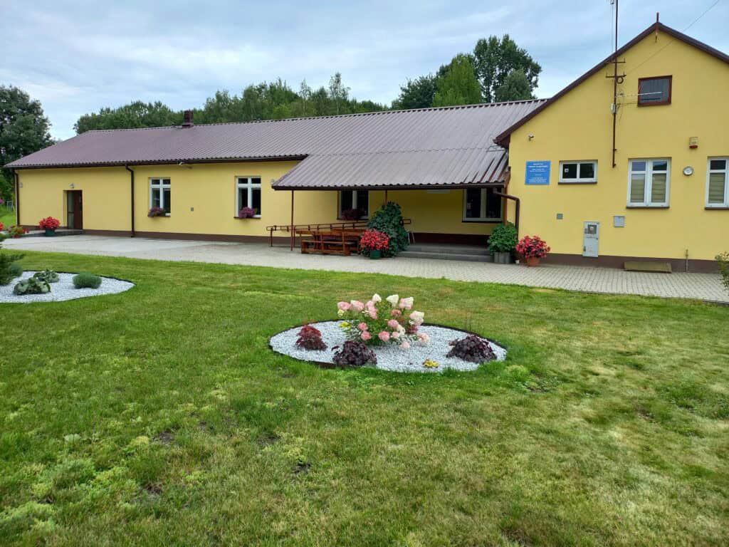 Żółty budynek z brązowym dachem, z kilkoma oknami ozdobionymi skrzynkami na kwiaty, stoi na bujnym, zielonym trawniku. Na pierwszym planie znajdują się rabaty kwiatowe z różowymi kwiatami, otoczone białym żwirem. Ścieżka prowadząca do wejścia jest wybrukowana, a na ganku widoczne są miejsca do siedzenia na zewnątrz.