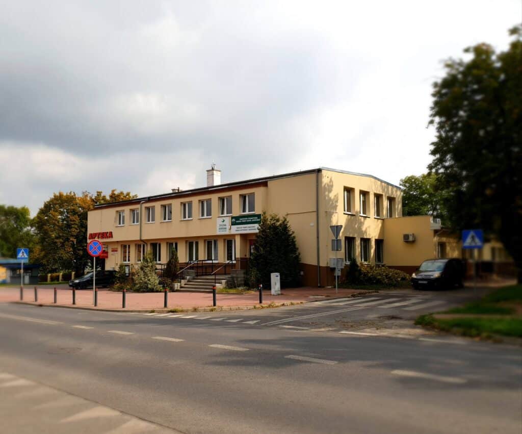 Piętrowy beżowy budynek z szyldem „Apteka”, położony na rogu ulicy z przejściami dla pieszych i otoczony drzewami.