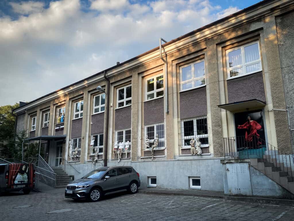 Dwukondygnacyjny budynek z dużymi oknami, posągami i manekinami przy zewnętrznych oknach, samochodem zaparkowanym przed budynkiem oraz zdjęciem w drzwiach przedstawiającym kobietę w czerwonej sukience.