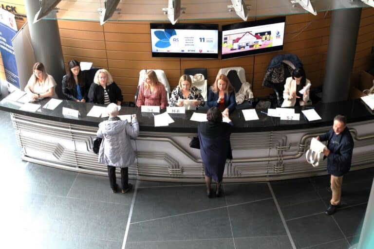 Widok „z góry” na punkt powitań i obsługi uczestników. Za kontuarem siedem pań witających gości. Przed kontuarem goście spotkania rejestrujący obecność (3 osoby)