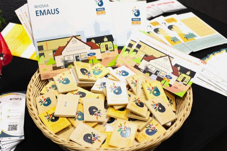 Ulotki i broszury ROPS i Wioski Emaus. W wiklinowym koszyku magnesy pamiątkowe z logo Emaus Lublin