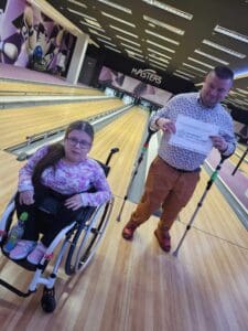 Przy torach do kręgli pozują kobieta na wózku inwalidzkim oraz mężczyzna poruszający się o dwóch kulach, który trzyma kartkę z logotypem Ministerstwa Sportu i Turystyki, dofinansowującego projekt