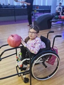 Kobieta na wózku inwalidzkim pozuje przy torze do kręgli. Przed nią stoi szyna prowadząca, na szczycie której umieszczono kulę do kręgli. Kobieta przytrzymuje ją opuszkami palców