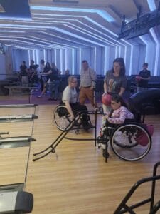Wnętrze kręgielni Masters Bowling & Bilard w Galerii Vivo. Uczestniczka na wózku inwalidzkim przygotowuje się do rzutu, ustawia rampę do kręgli. Asystuje jej stojąca obok kobieta, która trzyma kulę do strącania kręgli