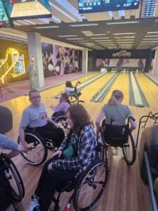 Sześciu uczestników rozgrywek na wózkach inwalidzkich stoi przy torach do kręgli, w tle widać częściowo zbite kręgle