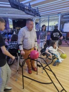 Uczestnik rozgrywek poruszający się o dwóch kulach korzysta z szyny prowadzącej, na której umieszczona jest kula do kręgli. Asystuje mu młodzieniec, a w tle widać graczy na sąsiednich torach