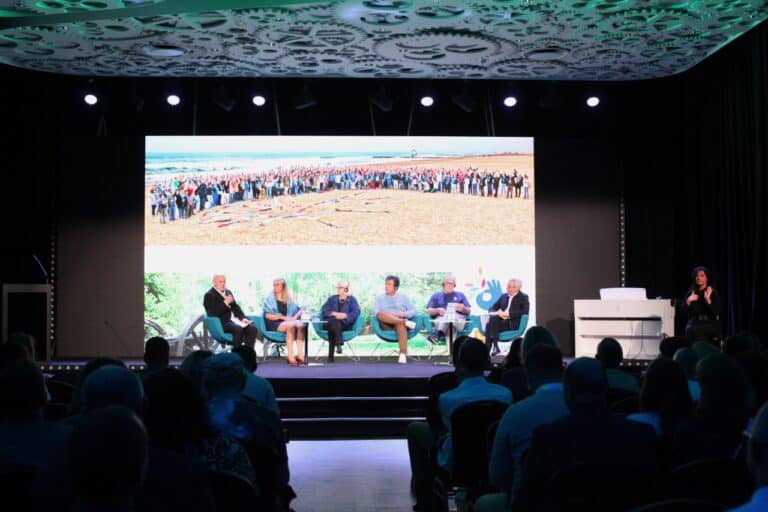 W centralnym punkcie kadru ekran, na tle którego w fotelach siedzą zaproszeni goście. Wśród nich są: osoby reprezentujące organizację Lekarze Bez Granic i współzałożyciele Emaus i Fundacji Między Nami, a także prof. Wojciech Otrębski. Rozmowę z nimi prowadzi Zbigniew Drążkowski. Po prawej stronie tłumaczka języka migowego