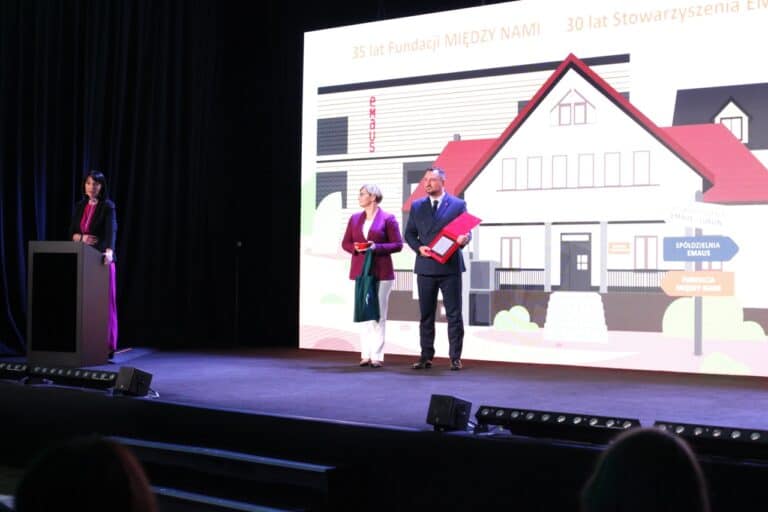 Widok na scenę. Od lewej, przy mównicy Iwona Kędziera. W centralnym punkcie kadru Wicemarszałek Województwa Lubelskiego Marek Wojciechowski prezentujący zgromadzonym przygotowane do wręczenia wyróżnienie