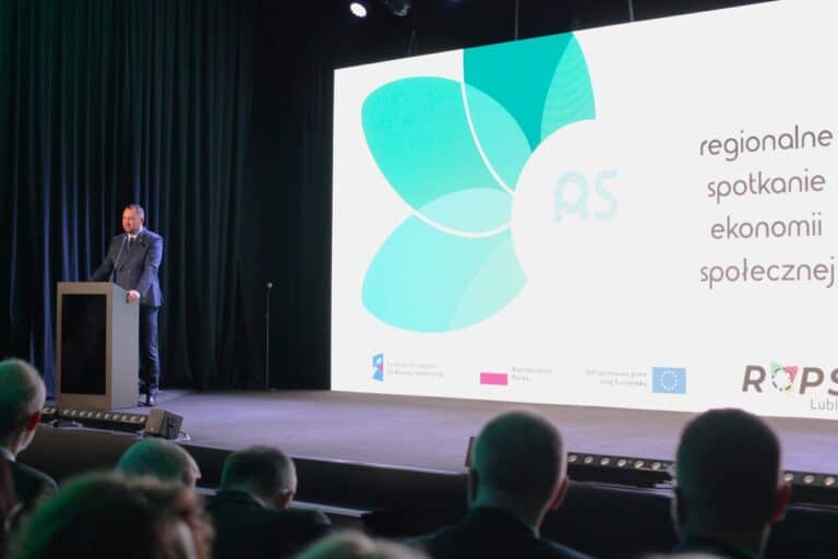 Widok na scenę. Po lewej stronie, przy mównicy Wicemarszałek Województwa Lubelskiego Marek Wojciechowski. W prawej i środkowej części kadru ekran z wyświetlonym napisem: Regionalne spotkanie ekonomii społecznej