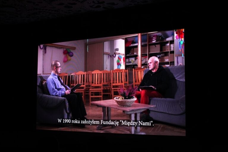 Widok na ekran z zatrzymanym w kadrze fragmentem wywiadu ze Zbigniewem Drążkowskim. Na pasku napisów, na dole ekranu wyświetla się: „W 1990 roku założyłem Fundację Między Nami”