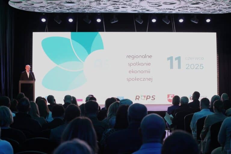 Fotografia zrobiona z perspektywy tylnej części sali. W centralnym miejscu w kadrze ekran z wyświetlonym napisem: „Regionalne spotkanie ekonomii społecznej. 11 czerwca, 2025”. Po lewej stronie, przy mównicy Zbigniew Drążkowski