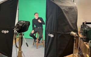 Przed ekranami jest tłumacz języka migowego, który wykonuje tłumaczenie przed kamerą siedząc na tle green screenu.