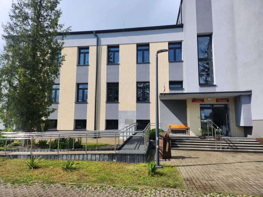 Jasny budynek z dużymi oknami, głównym wejściem ze schodami i rampą oraz niewielkim zagospodarowanym terenem z trawą i drzewem iglastym przed budynkiem.
