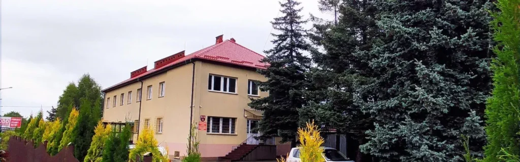 Budynek dwukondygnacyjny beżowy z czerwonym dachem, położony w otoczeniu iglastych drzew.