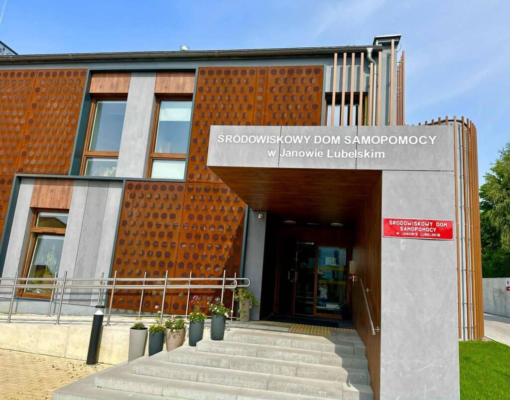 Budynek z szyldem nad wejściem: Środowiskowy Dom Samopomocy w Janowie Lubelskim, przy drzwiach rampa i doniczkowe rośliny.