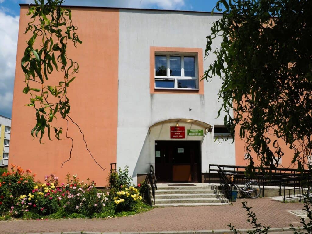 Dwukondygnacyjny budynek z brzoskwiniowymi i białymi ścianami, z łukowatym wejściem, schodami, rampą, klombami oraz zaparkowanym przy rampie rowerem.