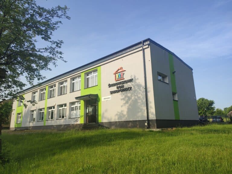 Dwukondygnacyjny budynek z zielonymi i beżowymi ścianami zewnętrznymi i szyldem na froncie, otoczony trawą i drzewami.