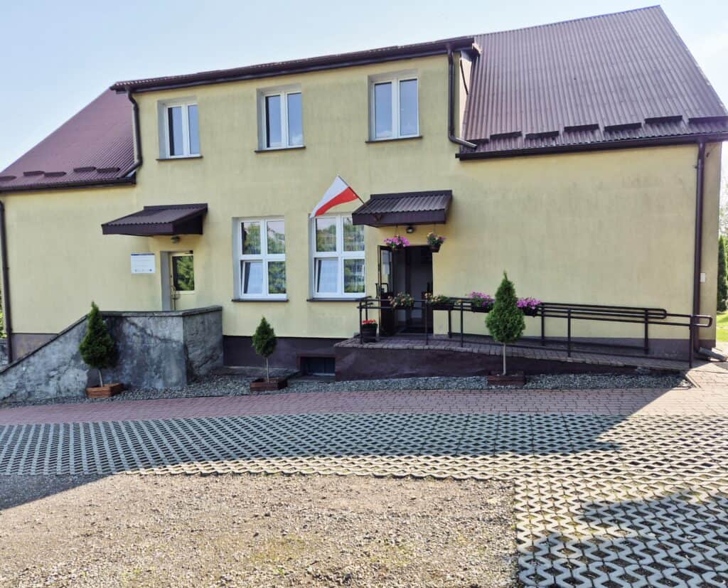 Dwukondygnacyjny żółty budynek z brązowym dachem, z podjazdem dla wózków inwalidzkich, roślinami doniczkowymi i polską flagą nad wejściem.