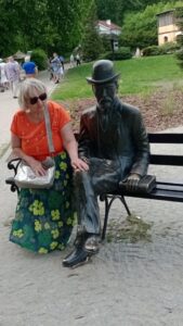 Kobieta siedzi na ławeczce obok rzeźby Bolesława Prusa. Postać Prusa jest odlana z brązu; pod jego lewą rękę znajduje się odlana również z brązu książka. Ławka znajduje się w przestrzeni publicznej, w tle widoczne drzewa i fragmenty budynków