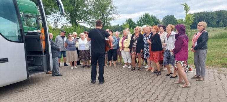 W plenerze, przed autokarem, stoi grupa wspólnie śpiewających seniorów z przewagą kobiet. Przed nimi, tyłem do fotografującego, stoi instruktor akompaniujący im na gitarze