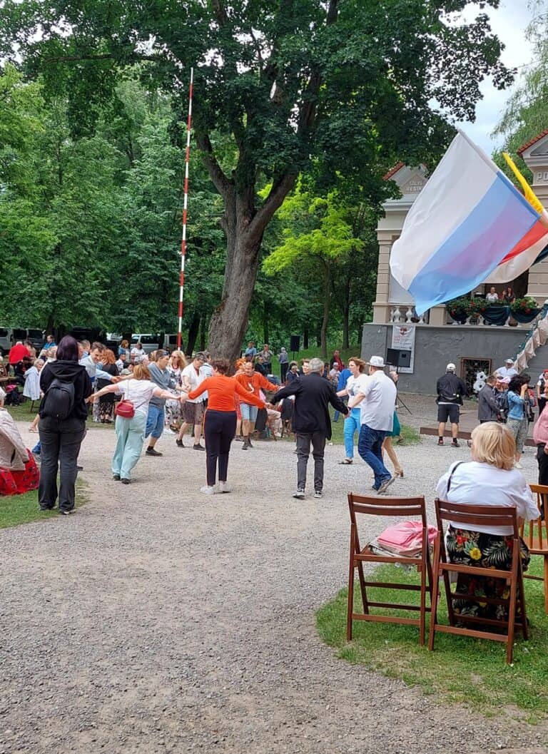 W plenerze grupa osób obu płci w różnym wieku tańczy w okręgu, trzymając się za ręce; w tle zielone drzewa