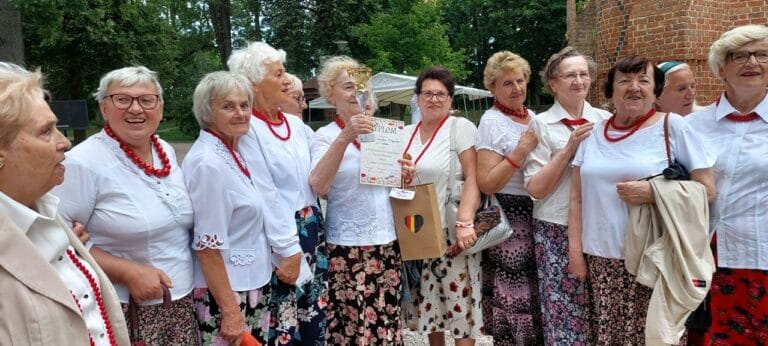 Grupa kobiet ubranych w białe bluzki, czerwone korale i kwieciste spódnice. Jedna z nich prezentuje puchar i dyplom otrzymane za występ podczas festiwalu