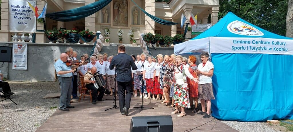 Grupa seniorek śpiewa na scenie, ubrane w stroje stylizowane na ludowe: białe bluzki, kwieciste spódnice i czerwone korale. Towarzyszą im seniorzy grający na skrzypcach, gitarach i akordeonie. W tle widoczny jest budynek z wysoką podmurówką, ozdobiony flagami w barwach: biało-czerwonej, biało-niebieskiej i biało-żółtej. Po lewej stronie wisi baner z napisem „Nadbużański Integracyjny Festiwal Muzyczny, Kodeń”, a po prawej znajduje się duży niebieski namiot z napisem „Gminne Centrum Kultury, Sportu i Turystyki w Kodniu”.