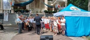 Grupa seniorek śpiewa na scenie, ubrane w stroje stylizowane na ludowe: białe bluzki, kwieciste spódnice i czerwone korale. Towarzyszą im seniorzy grający na skrzypcach, gitarach i akordeonie. W tle widoczny jest budynek z wysoką podmurówką, ozdobiony flagami w barwach: biało-czerwonej, biało-niebieskiej i biało-żółtej. Po lewej stronie wisi baner z napisem „Nadbużański Integracyjny Festiwal Muzyczny, Kodeń”, a po prawej znajduje się duży niebieski namiot z napisem „Gminne Centrum Kultury, Sportu i Turystyki w Kodniu”.