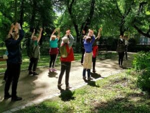 Grupa seniorek bierze udział w ćwiczeniach rozciągających na świeżym powietrzu, unosząc ramiona nad głowę w otoczeniu zalesionego parku