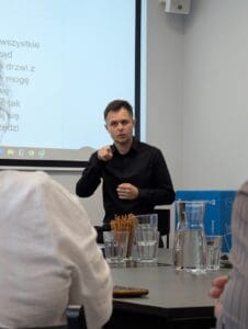 Mężczyzna w czarnej koszuli stoi przed ekranem projekcyjnym i tłumaczy wypowiedź w języku migowym, wykonując gest palcem, skierowany w stronę widowni, na znak „Ty”. Na ekranie widoczny jest fragment napisów na żywo w języku polskim. Na pierwszym planie widać uczestników spotkania siedzących przy stole, szklanki z wodą, dzbanek i pojemnik z paluszkami. W tle po prawej stronie znajduje się niebieskie pudełko z napisem „Lublin is Youth”