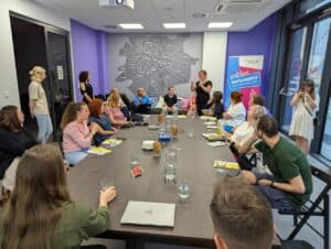 Uczestnicy spotkania siedzą wokół dużego stołu w nowoczesnej sali z fioletowymi akcentami. Na pierwszym planie widać laptopy, szklanki z wodą, broszury z symbolem dostępności, talerzyk z cukierkami i paluszkami. W tle kobieta w czerni tłumaczy wypowiedź w języku migowym, a pozostali obecni słuchają z zainteresowaniem. Na ścianie znajduje się duża mapa Lublina, a po prawej stronie baner z napisem „Miasto Młodych - Polityka Młodzieżowa Lublina”. Jedna z uczestniczek robi zdjęcie telefonem