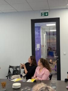 Dwie kobiety siedzą przy stole w sali spotkań i rozmawiają. Kobieta po lewej w ciemnych włosach i czarnych okularach, ubrana na czarno, wypowiada się w PJM. Kobieta obok niej ma długie kręcone włosy, upięte w kucyk, w różowej koszuli w białe paski, patrzy w jej stronę z uwagą. Na stole leżą broszury z symbolem dostępności, szklanki z wodą, smartfon, ciastka oraz talerzyk z paluszkami. W tle przeszklone drzwi z oznaczeniem „03” i widoczną częścią przestrzeni z leżakami oraz napisem „Urban Lab”. Nad drzwiami mieści się znak wyjścia ewakuacyjnego