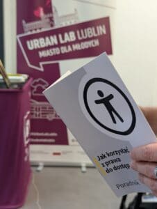 Zbliżenie na męską dłoń trzymającą broszurę z napisem „Jak korzystać z prawa do dostępności? Poradnik” oraz symbolem dostępności – ilustracją-ikonką człowieka w czarnym okręgu. W tle widoczny baner w kolorze fioletowym z napisem „Urban Lab Lublin - Miasto dla Młodych” oraz stylizowaną grafiką budynku. Baner stoi na tle jasnej ściany, obok stolika z innymi materiałami informacyjnymi