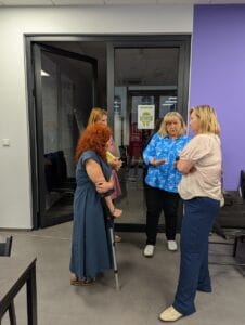 Grupa czterech kobiet prowadzi rozmowę w przejściu przy przeszklonych drzwiach. Jedna z kobiet, z rudymi kręconymi włosami, opiera się na kuli ortopedycznej. Inna, ubrana w niebieską koszulę z białymi wzorami, gestykuluje podczas mówienia. Pozostałe uczestniczki rozmowy słuchają uważnie. W tle na szybie widoczna jest kartka z napisem „Zielone Sieci” i kodem QR. Ściany są w odcieniach bieli i fioletu, a podłoga wykonana jest z szarego materiału