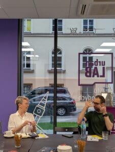 Dwie osoby siedzą przy stole w jasnym pomieszczeniu z dużymi oknami. Kobieta po lewej, w białej koszuli, trzyma zapakowane ciastko w dłoniach i spogląda w stronę rozmówcy uśmiechając się. Mężczyzna po prawej, w zielonej koszulce, pokazuje znaki w języku migowym. Na stole leżą filiżanki, szklanki z wodą, talerzyki, paluszki, smartfony i książeczki z symbolem dostępności. Przez okno widać zaparkowany samochód i fasadę budynku naprzeciwko. Na szybie znajduje się logo „Urban Lab”