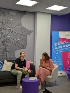 Dwie kobiety prowadzą rozmowę na tle szarej ściany, na której znajduje się duża mapa dzielnic Lublina na ścianie oraz baner z napisem „Miasto Młodych – Polityka Młodzieżowa Lublina”. Jedna z kobiet, ubrana w czarną bluzkę i jasne spodnie, siedzi na ciemnej kanapie ozdobionej kolorowymi poduszkami. Druga, w różowej sukience w drobny wzór, siedzi na jasnym fotelu. Między nimi stoi niski, fioletowy stolik z kartką i szklanką wody